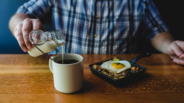 Man pouring cream into coffee, skillet of hash with egg on top